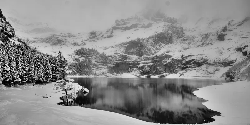 Black and white landscape of a frozen alpine lake surrounded by snow-covered mountains and evergreen forests under overcast skies.